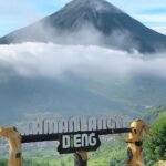 Mountain peak rising above clouds, with a scenic overlook featuring carved wooden goats and a Dieng sign in the foreground