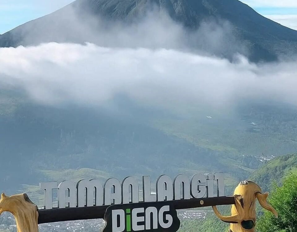 Mountain peak rising above clouds, with a scenic overlook featuring carved wooden goats and a Dieng sign in the foreground