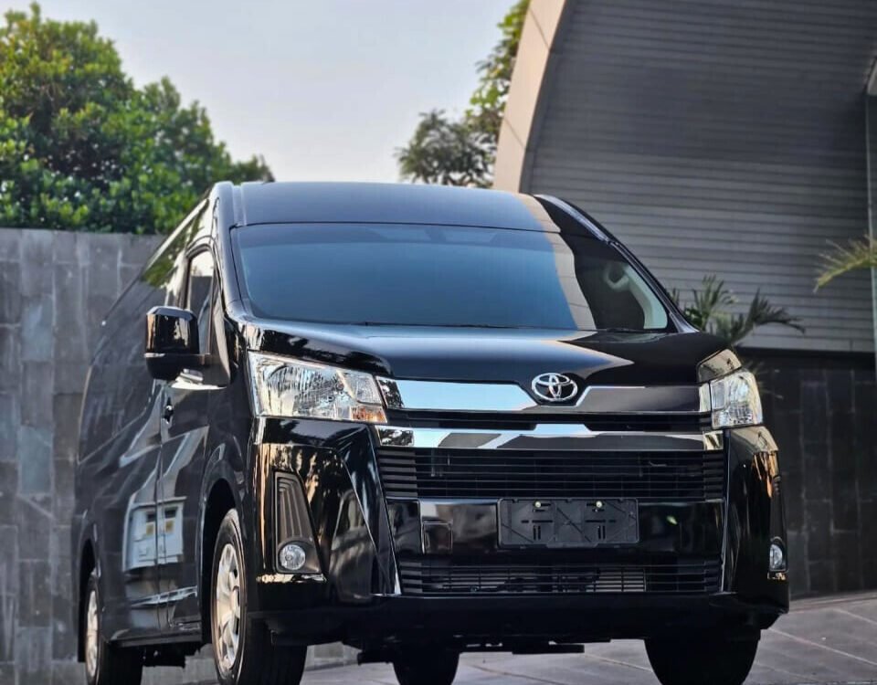 premio htm nasmoco edit - Dyantransport Front view of a black Toyota minivan parked on a paved surface near a modern building.