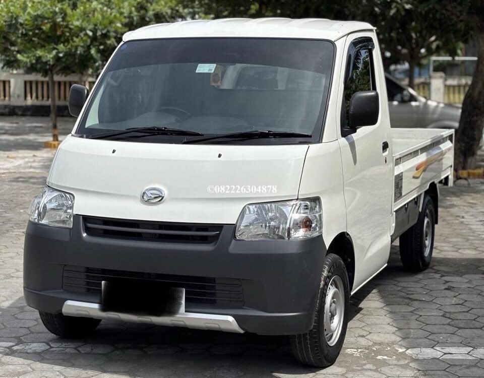 White compact pickup truck parked on a stone-paved lot with green trees in the background (front-left view).