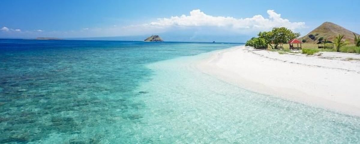 Tropical beach with clear turquoise water, white sand, and a small gazebo on the right under a blue sky.