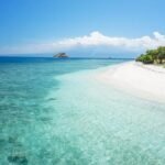 Tropical beach with clear turquoise water, white sand, and a small gazebo on the right under a blue sky.