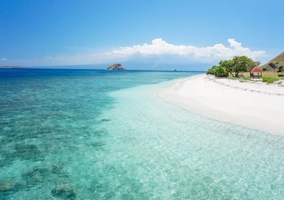 Tropical beach with clear turquoise water, white sand, and a small gazebo on the right under a blue sky.