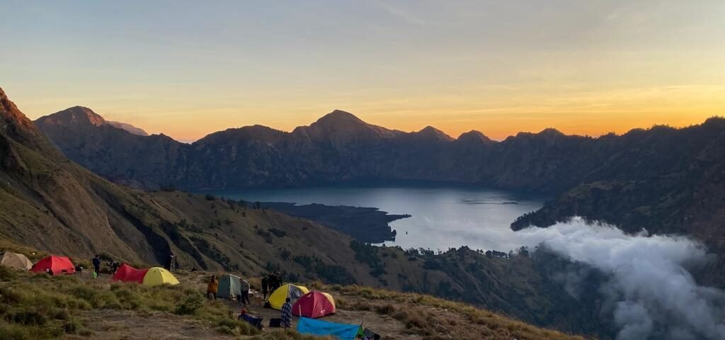 Campsite on a grassy mountainside overlooking a lake at sunset, colorful tents scattered among hikers and gear with rugged peaks in the distance.