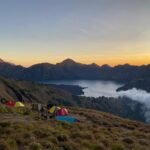 Campsite on a grassy mountainside overlooking a lake at sunset, colorful tents scattered among hikers and gear with rugged peaks in the distance.