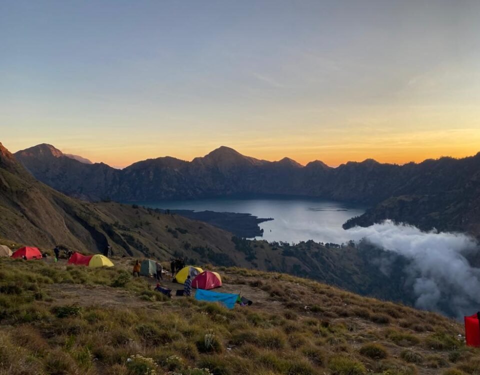Campsite on a grassy mountainside overlooking a lake at sunset, colorful tents scattered among hikers and gear with rugged peaks in the distance.