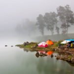 Misty lakeside campsite with colorful tents on a rocky shore and calm water reflecting the scene