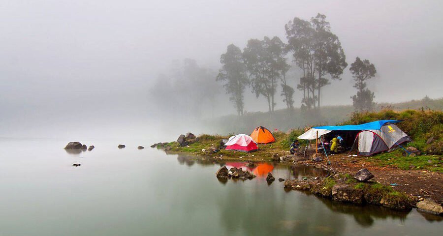 Misty lakeside campsite with colorful tents on a rocky shore and calm water reflecting the scene