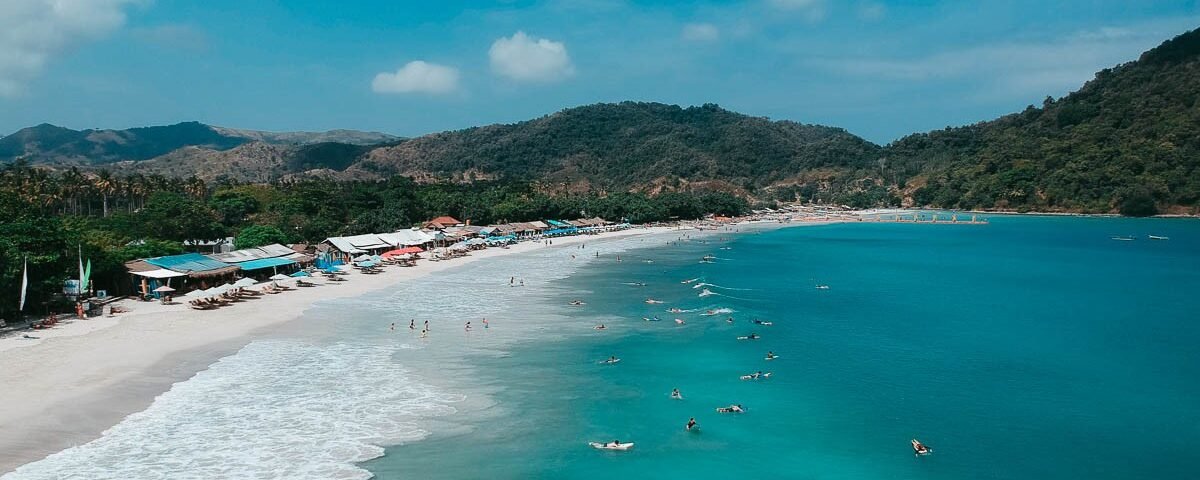 Sunny tropical beach with white sand, turquoise water, row of umbrellas, and forested hills in the background.
