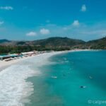 Sunny tropical beach with white sand, turquoise water, row of umbrellas, and forested hills in the background.