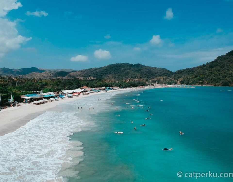 Sunny tropical beach with white sand, turquoise water, row of umbrellas, and forested hills in the background.