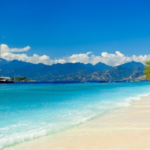 Turquoise water and white sandy beach with a sailboat near shore and green trees on the right, distant mountains under a blue sky.