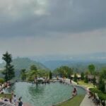 Circular infinity pool on a hillside resort, people lounging and swimming with a green valley and cloudy sky in the distance
