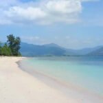 Sunlit white sandy beach with turquoise water and distant mountains under a blue sky.