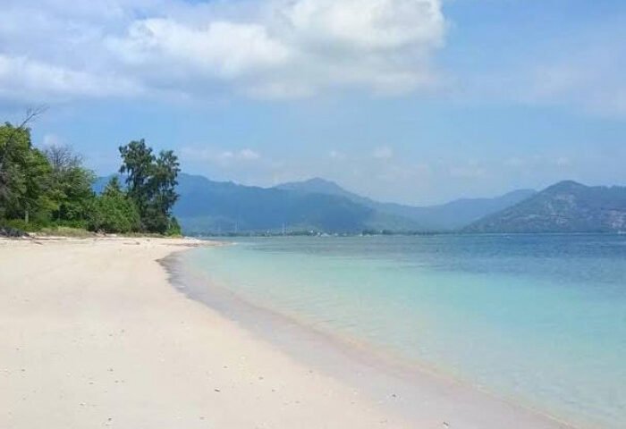 Sunlit white sandy beach with turquoise water and distant mountains under a blue sky.