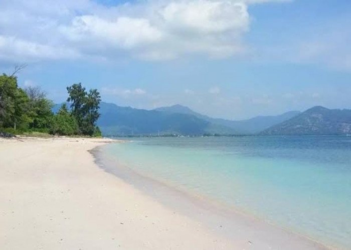 Sunlit white sandy beach with turquoise water and distant mountains under a blue sky.