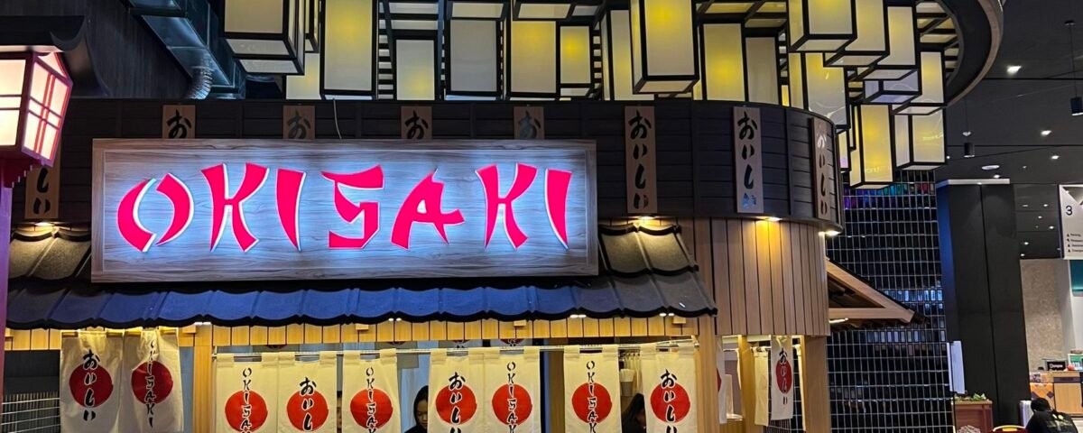 Curved neon sign reading "Welcome to Harajuku Street" above a Japanese restaurant counter in a mall.