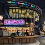 Curved neon sign reading "Welcome to Harajuku Street" above a Japanese restaurant counter in a mall.