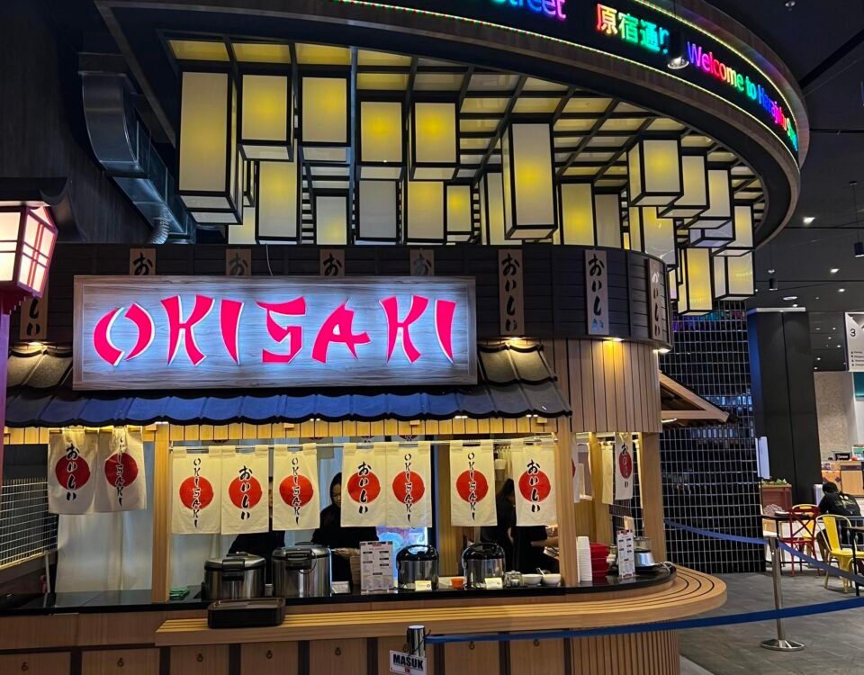 Curved neon sign reading "Welcome to Harajuku Street" above a Japanese restaurant counter in a mall.