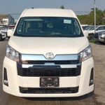 Front view of a white Toyota minivan parked in a car dealership lot with other vehicles in the background.
