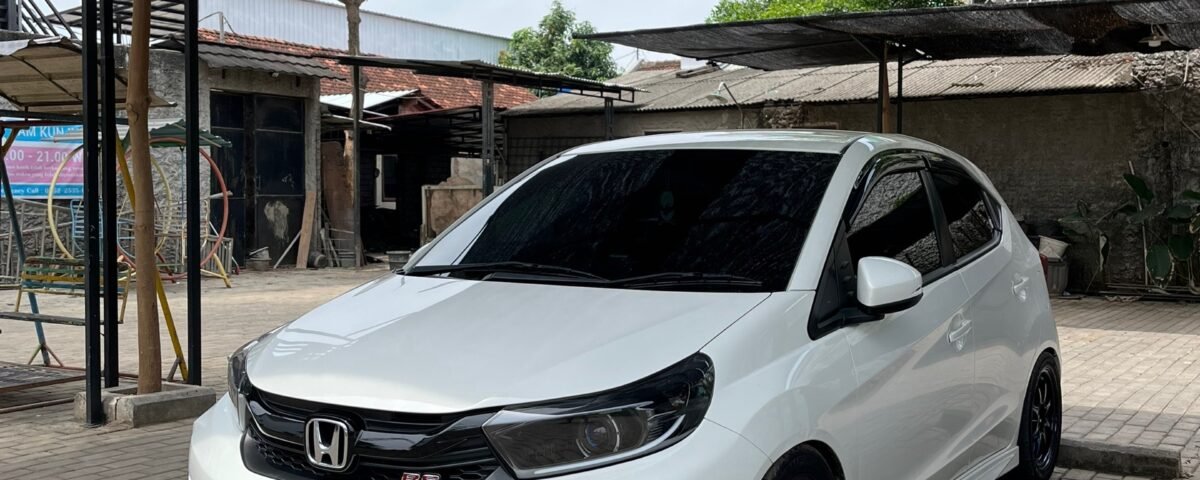 White Honda RS hatchback parked on brick pavers under dark shade sails in an outdoor lot with buildings in the background.