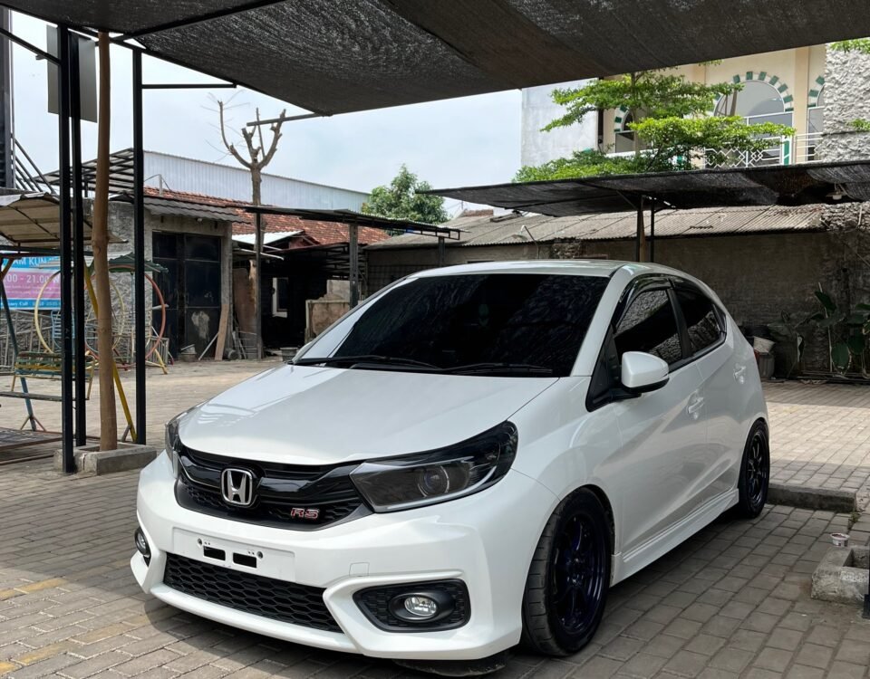 White Honda RS hatchback parked on brick pavers under dark shade sails in an outdoor lot with buildings in the background.