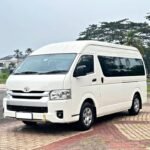 snaptik_7500188955992804614_2_v2 - Dyantransport White Toyota van with a high roof parked on a brick-paved lot, with trees in the background.