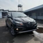 Black Toyota SUV parked in an industrial parking lot, seen from the front three-quarter angle with a tall metal watchtower in the background.