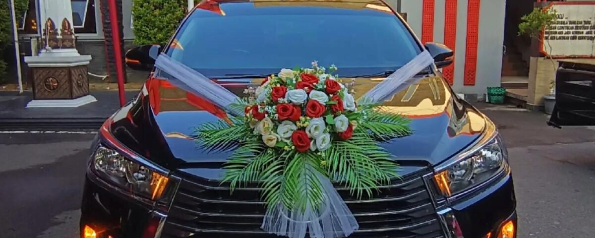 Black wedding car with a large red/white flower bouquet on the hood and white ribbons, parked in front of a monument with a crest on a stone wall behind it.