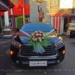 Black wedding car with a large red/white flower bouquet on the hood and white ribbons, parked in front of a monument with a crest on a stone wall behind it.
