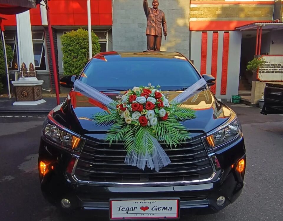 Black wedding car with a large red/white flower bouquet on the hood and white ribbons, parked in front of a monument with a crest on a stone wall behind it.