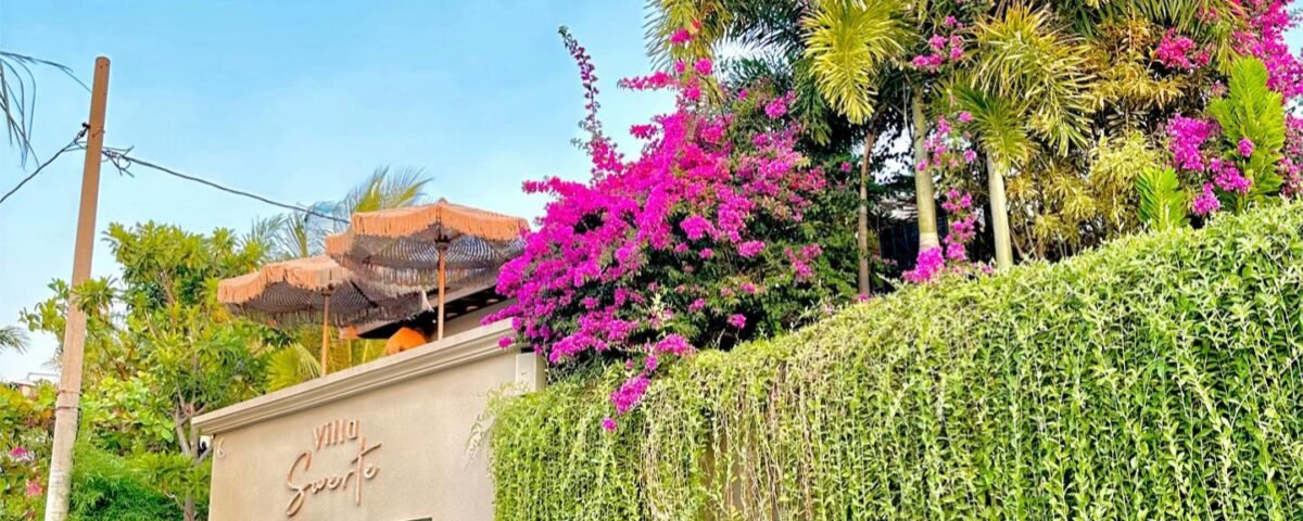 Cream villa wall with a lush green vine hedge and pink bougainvillea, under a bright blue sky (for sale sign visible).