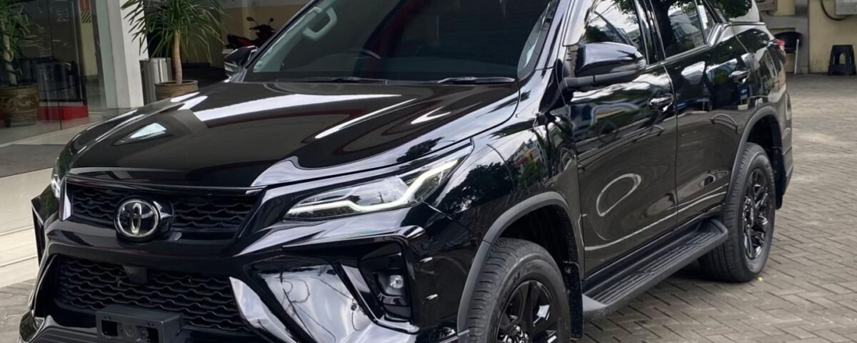 Black Toyota SUV (Fortuner) parked on a brick-paved driveway beside a modern building with glass doors and red structural beams, front three-quarter view.