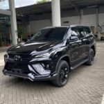 Black Toyota SUV (Fortuner) parked on a brick-paved driveway beside a modern building with glass doors and red structural beams, front three-quarter view.