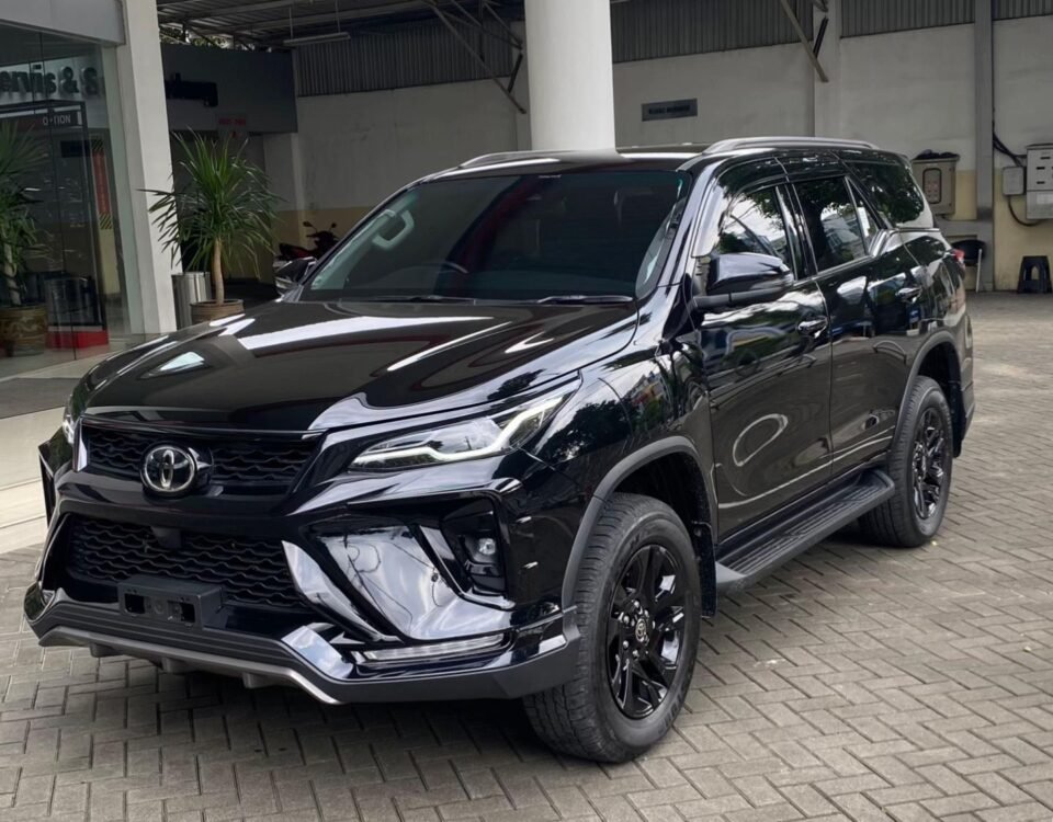 Black Toyota SUV (Fortuner) parked on a brick-paved driveway beside a modern building with glass doors and red structural beams, front three-quarter view.