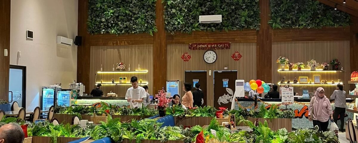 Interior of a restaurant with a tall wooden ceiling, warm lighting, and a lush green wall behind the counter; customers seated and dining nearby.