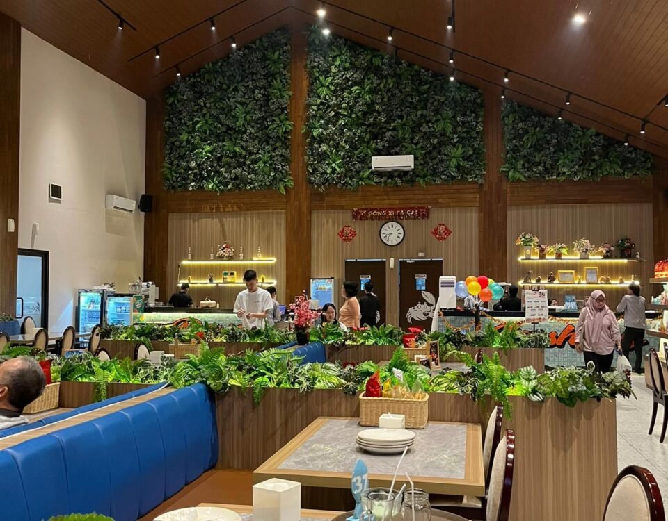 Interior of a restaurant with a tall wooden ceiling, warm lighting, and a lush green wall behind the counter; customers seated and dining nearby.