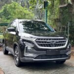Front three-quarter view of a dark gray SUV parked on a stone driveway with lush green foliage in the background, reflecting on the car’s surface.