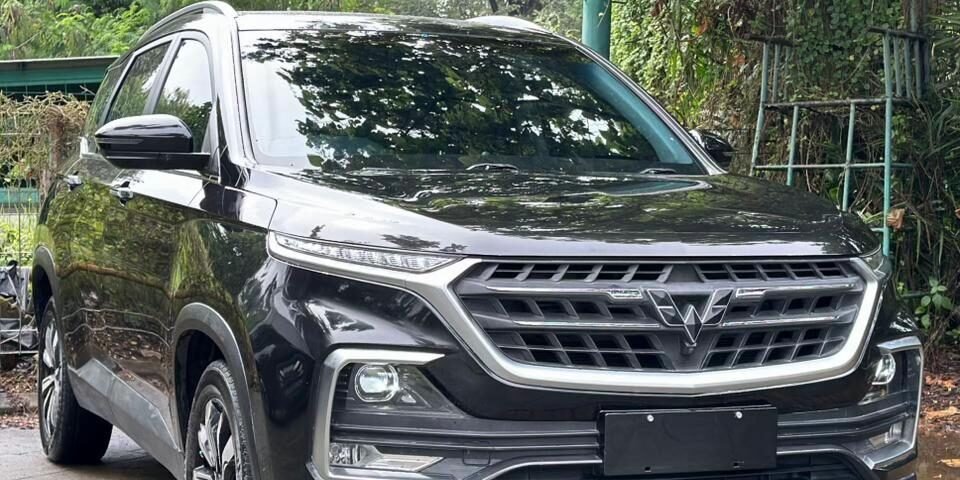 Front three-quarter view of a dark gray SUV parked on a stone driveway with lush green foliage in the background, reflecting on the car’s surface.