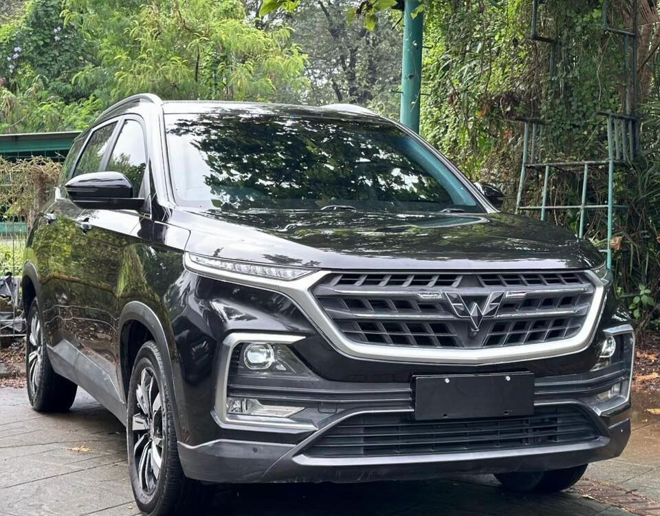 Front three-quarter view of a dark gray SUV parked on a stone driveway with lush green foliage in the background, reflecting on the car’s surface.