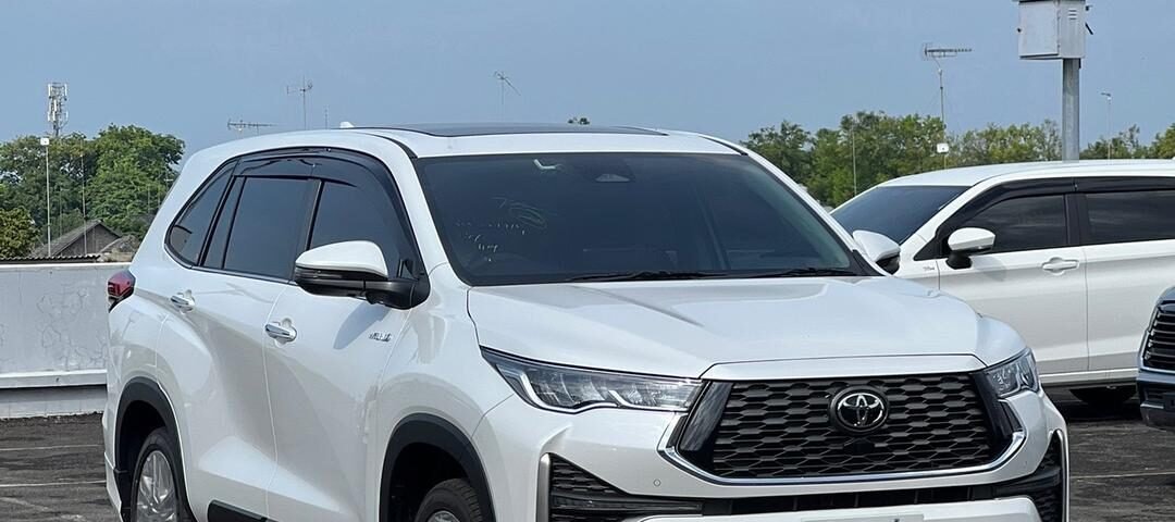 Front three-quarter view of a white Toyota SUV (Highlander) parked in an outdoor lot with other vehicles and blue sky behind it.