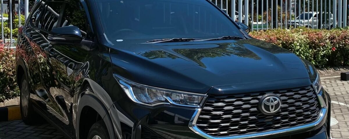 Front view of a dark Toyota SUV parked in a paved lot with a blue parking sign and greenery in the background under a clear blue sky.