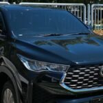 Front view of a dark Toyota SUV parked in a paved lot with a blue parking sign and greenery in the background under a clear blue sky.