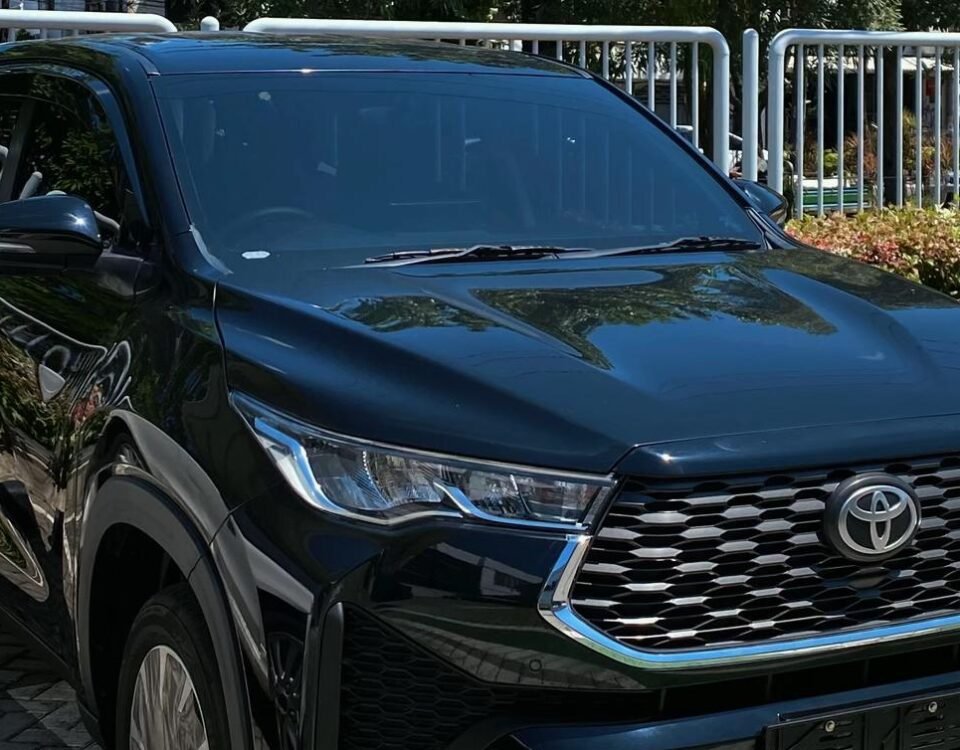 Front view of a dark Toyota SUV parked in a paved lot with a blue parking sign and greenery in the background under a clear blue sky.