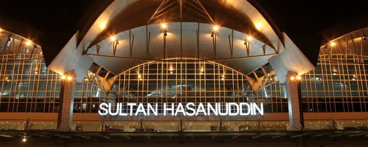 Night exterior of Sultan Hasanuddin Airport with a glass facade and the illuminated sign 'SULTAN HASANUDDIN' under a curved roof.