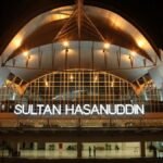 Night exterior of Sultan Hasanuddin Airport with a glass facade and the illuminated sign 'SULTAN HASANUDDIN' under a curved roof.