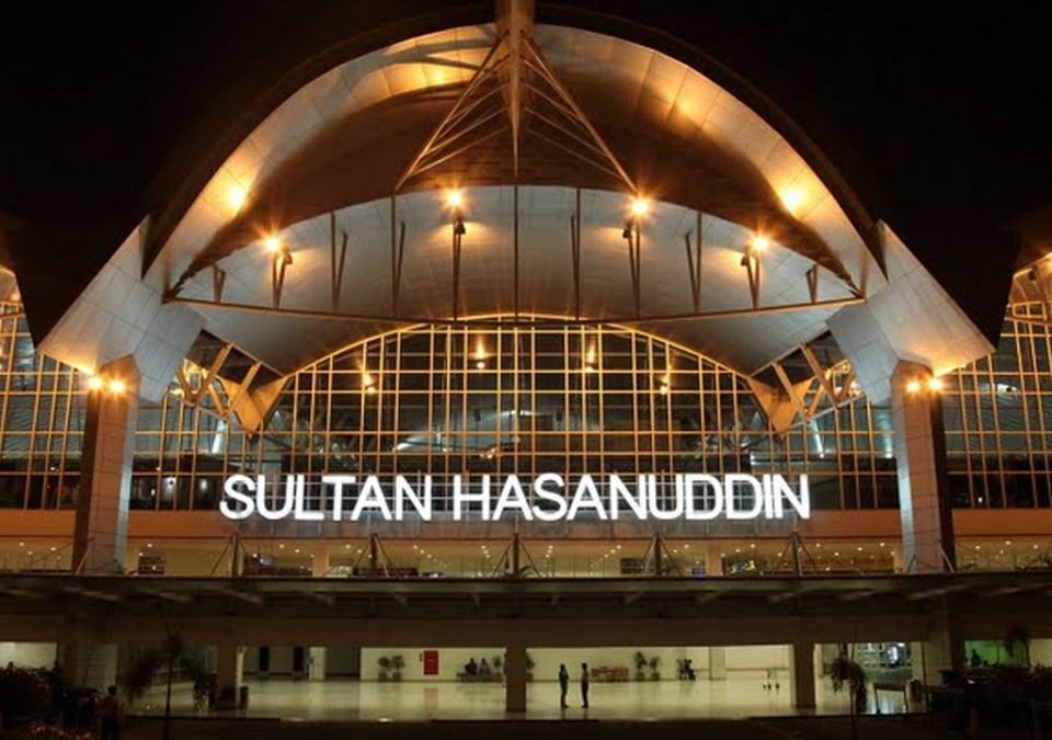 Night exterior of Sultan Hasanuddin Airport with a glass facade and the illuminated sign 'SULTAN HASANUDDIN' under a curved roof.