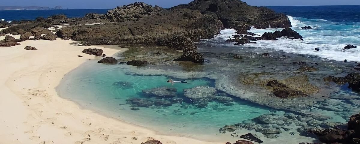 Turquoise tidal pool bordered by white sand and dark rocks, with a person snorkeling inside.