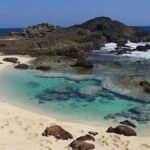 Turquoise tidal pool bordered by white sand and dark rocks, with a person snorkeling inside.