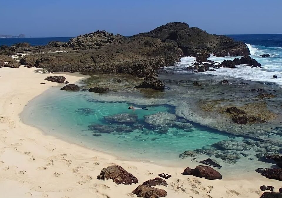 Turquoise tidal pool bordered by white sand and dark rocks, with a person snorkeling inside.
